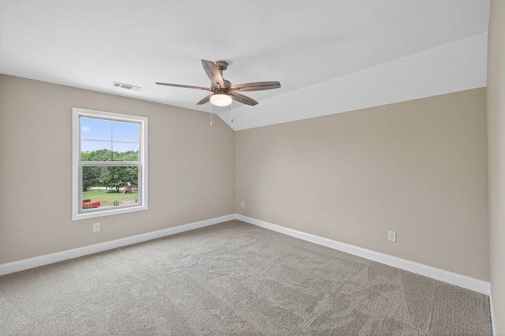 Carpeted room with white walls, ceiling fan with light fixture, large window overlooking playground and trees, crown molding along ceiling.