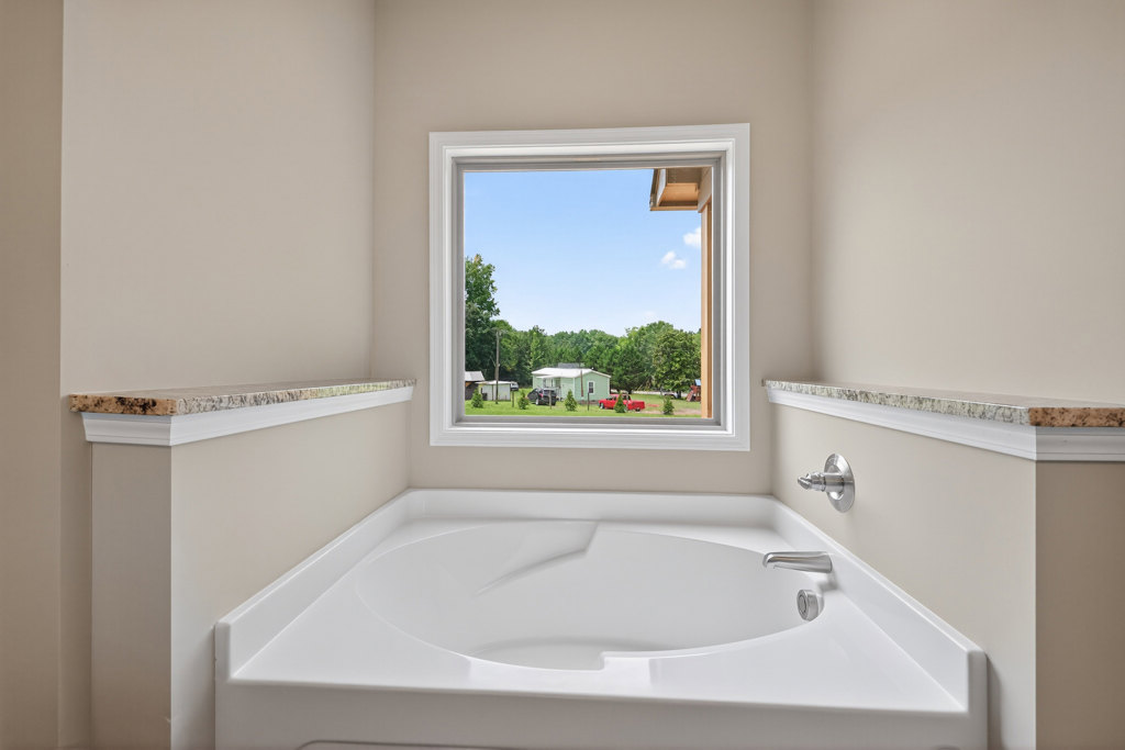 White freestanding bathtub with curved edge beside large window overlooking yard and trees, marble-topped white shelf and chrome hook visible in modern bathroom.