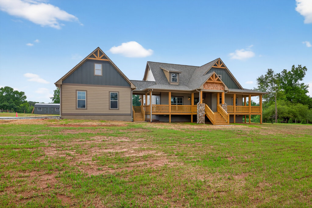 White farmhouse with covered front porch, triangular roofline, attached garage, expansive green lawn, mature tree, and blue sky overhead