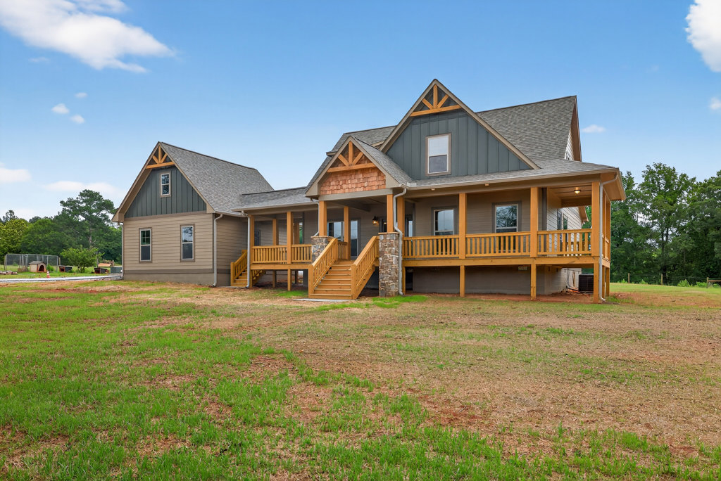 Two-story home with covered porch, stone retaining wall, wooden stairs leading to grassy lawn, large windows, and gabled roof under partly cloudy sky
