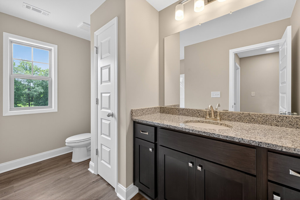 Bathroom with marble countertop, undermount sink, silver faucet, white toilet, white cabinetry, tile flooring, window showing trees, white door with silver knob