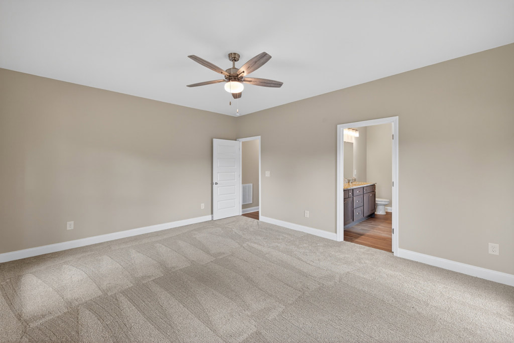 Carpeted bedroom with white walls, ceiling fan with light, white door featuring silver knobs, partial view into bathroom with toilet and sink, close-up of countertop