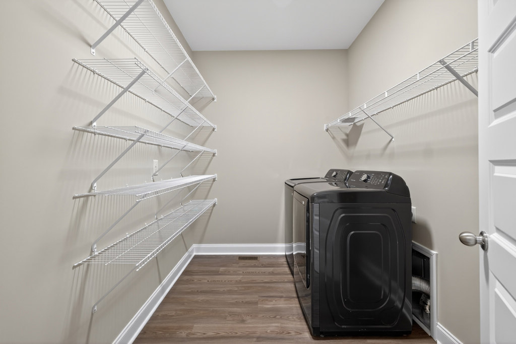 Front-loading black dryer on light wood floor, white metal shelf with railings mounted on plaster wall, white baseboard trim along floor, laundry room interior with neutral