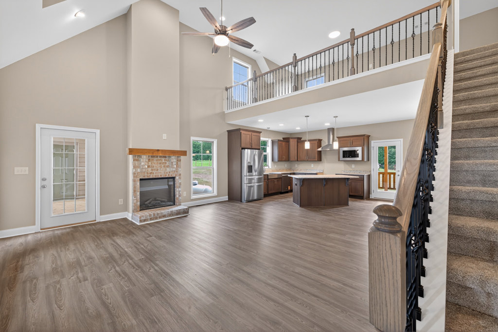 Spacious open floor plan featuring hardwood flooring, modern kitchen with white cabinetry, glass-paneled door, ceiling fan with light, fireplace with glass door, staircase with