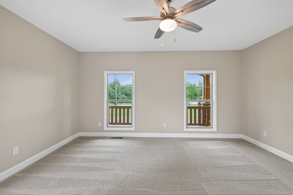 Carpeted room with white walls, ceiling fan with light fixture, large windows overlooking green trees and grass, wooden porch railing visible through window