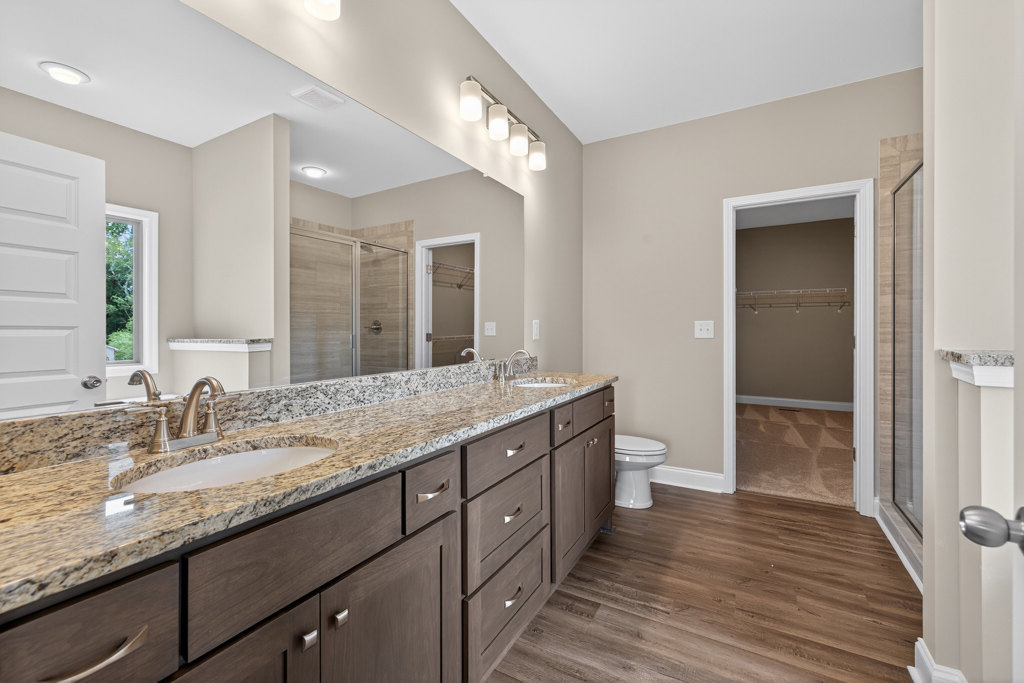 Bathroom featuring a marble countertop with undermount sink, large wall mirror, glass-enclosed shower, white toilet, and adjacent closet with shelving