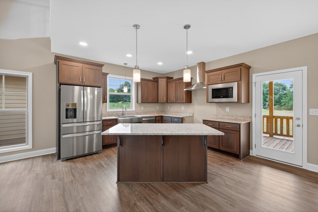Spacious kitchen featuring a large quartz island, stainless steel refrigerator and microwave, white cabinetry, glass-paneled door, and wide window above the sink