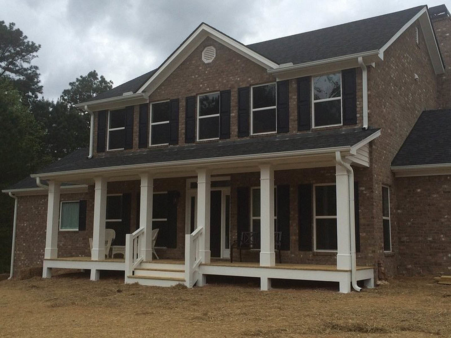 Spacious porch with white railings and steps, black-framed and white-framed windows, light siding, manicured lawn, leafy trees, and partly cloudy sky