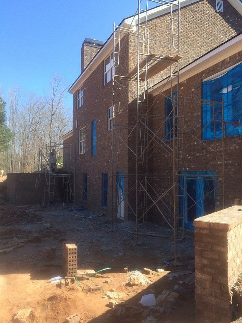 Scaffolding surrounds a partially built brick home exterior, blue tarp covers a window opening, white construction materials rest on the ground, trees and sky visible in