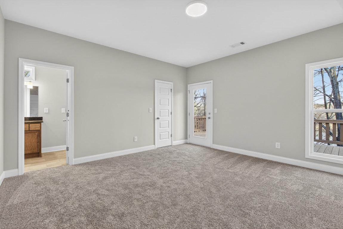 Beige carpeted room with white walls, white ceiling light, two white doors with silver knobs, window showing trees outside, and a railing near the entry.