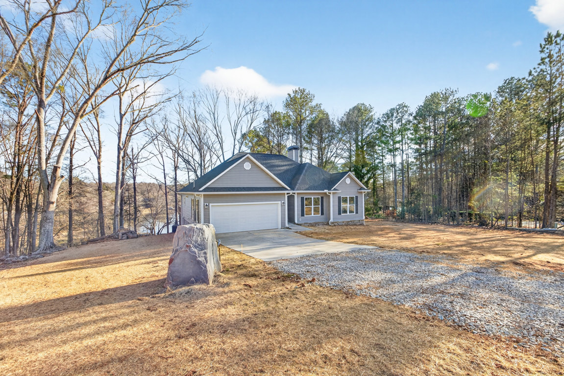 Two-story house with attached garage, gravel driveway, large rock near entrance, snow-covered ground, and mature trees in the background