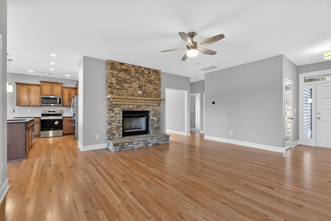 Living room with hardwood floors, stone step, glass-door fireplace, ceiling fan with light, partial view of kitchen oven and cabinetry