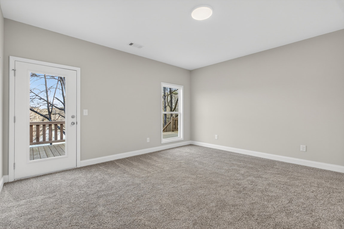 Carpeted room with white ceiling light, white door opening to wooden deck, tree and lake visible through glass window and railing