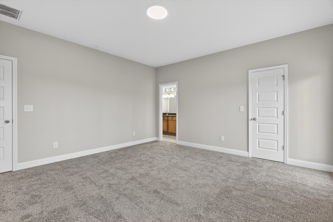 Carpeted bedroom with open white door, silver knob, white walls, bathroom vanity and mirror visible through doorway, ceiling light fixture, baseboard molding