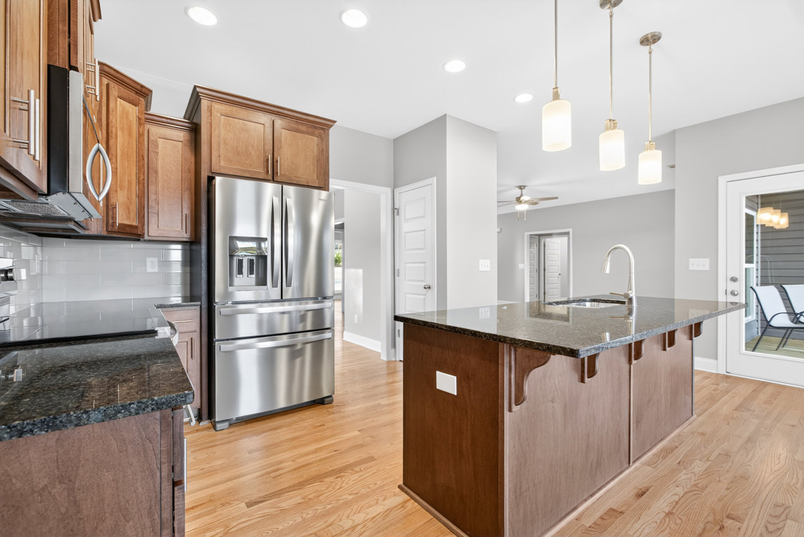 Stainless steel refrigerator beside wood cabinets in a kitchen with stone countertops, central island with sink, and pendant ceiling light