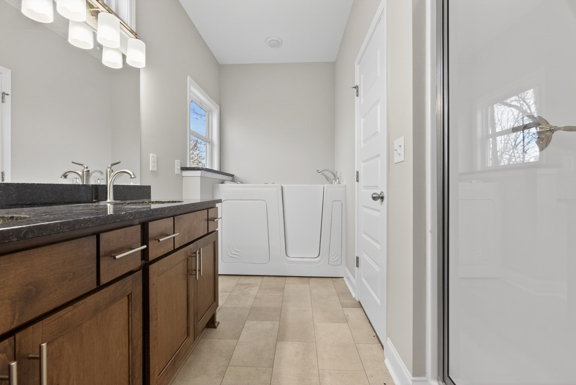 Modern bathroom featuring a freestanding white bathtub, rectangular sink on a stone countertop, glass shower door, light gray tile flooring, white cabinetry, and a window with blue