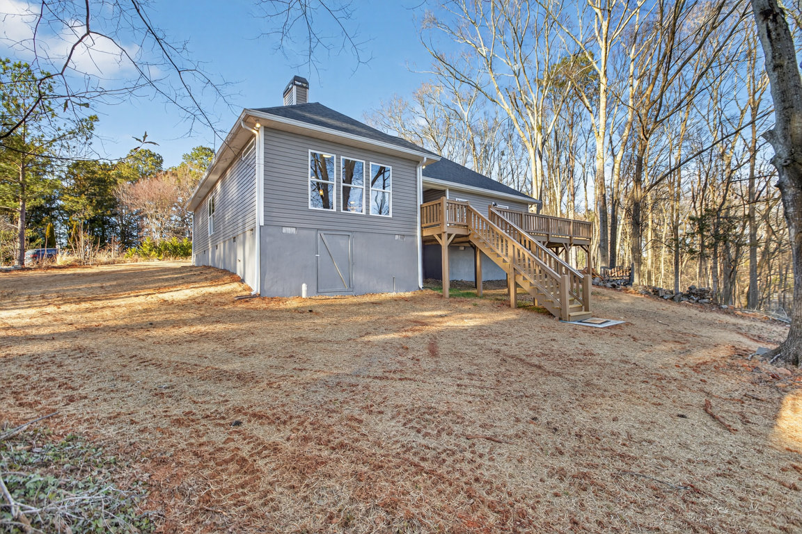Grey house with row of windows, wooden deck and staircase, metal door, chimney, surrounded by trees and plants.