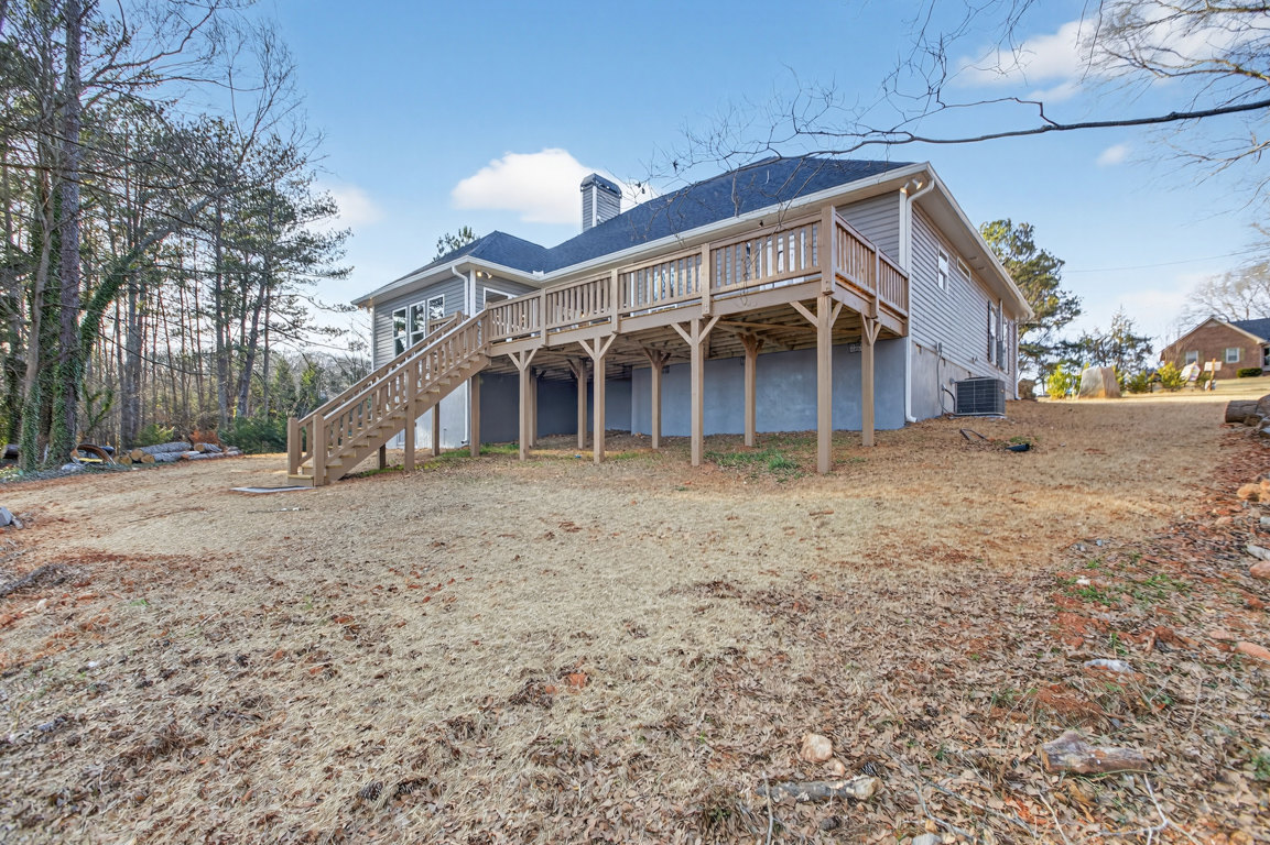 Two-story home with light siding, elevated wooden deck, outdoor stairs, and spacious yard covered in brown leaves, bordered by mature trees under a cloudy sky