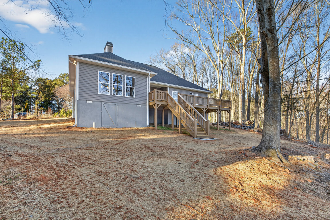 Two-story house with gray siding, row of large windows, wooden deck and staircase leading to grassy backyard, mature tree in the background, cloudy sky overhead