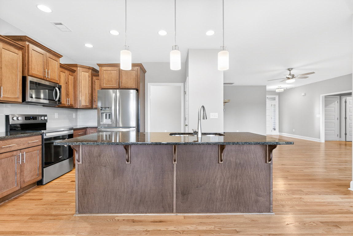 Spacious kitchen featuring a large central island with stone countertop, built-in sink, stainless steel refrigerator with open door, modern microwave, pendant light fixture, and