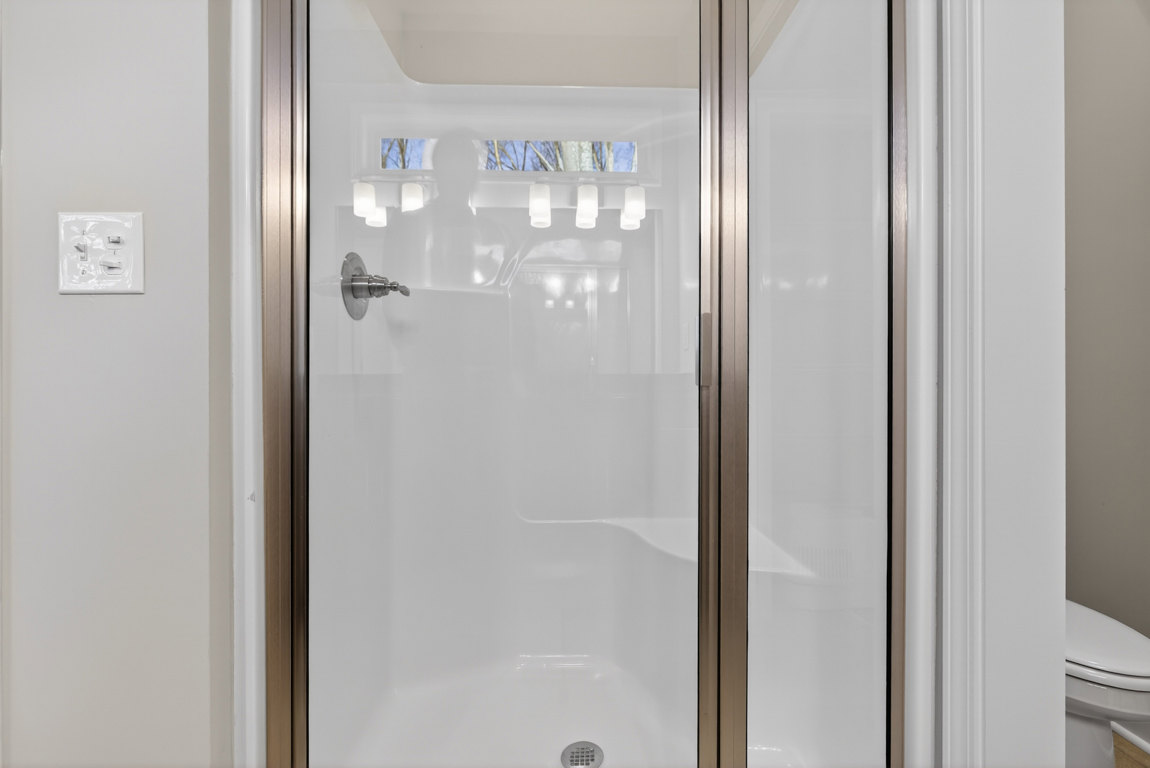 Walk-in shower with clear glass door, chrome faucet, white tile walls, nearby toilet, wall-mounted light fixture, skylight overhead, and reflection of person in glass
