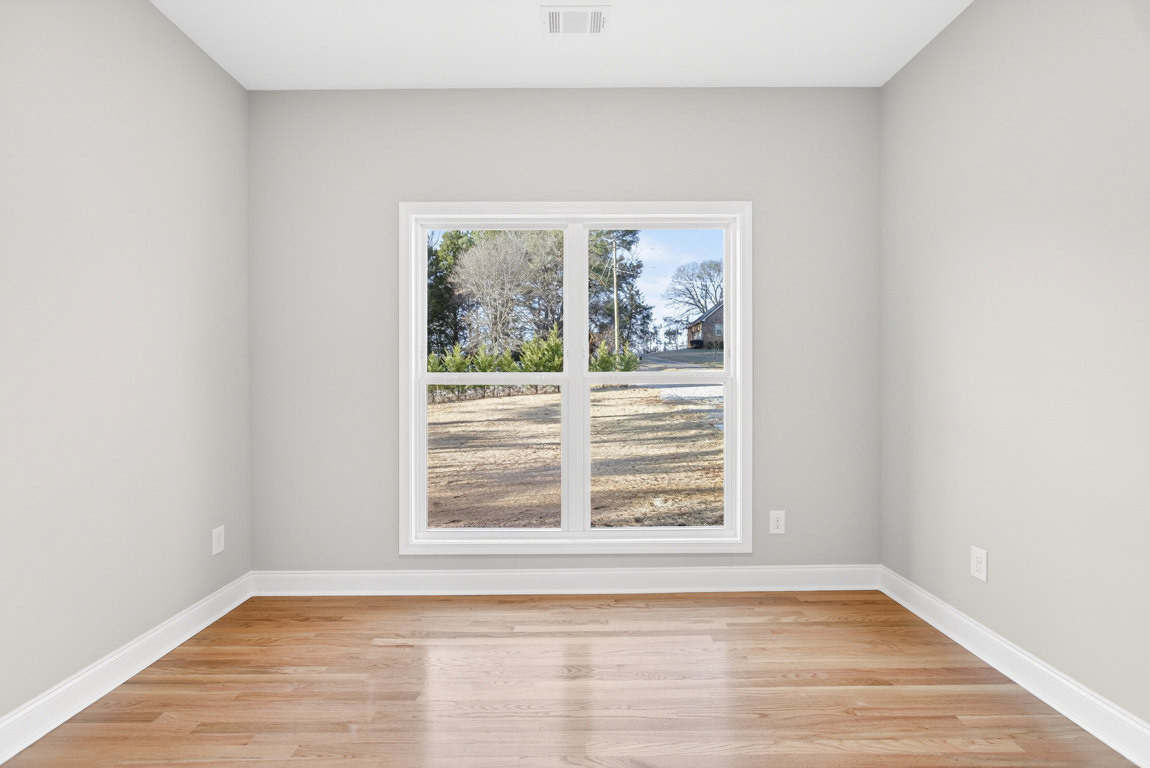 Sunlit room featuring wide-plank hardwood floors, white baseboards, and a large window overlooking trees and a dirt field.