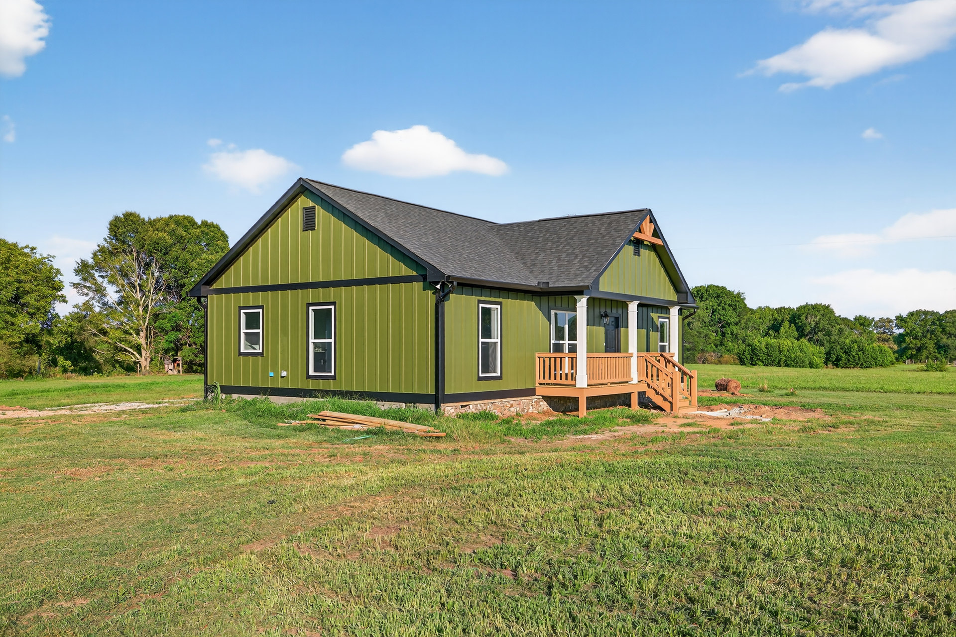 Green-painted farmhouse with yellow trim, white-framed windows, covered porch with railing, expansive grassy lawn, blue sky, and scattered trees