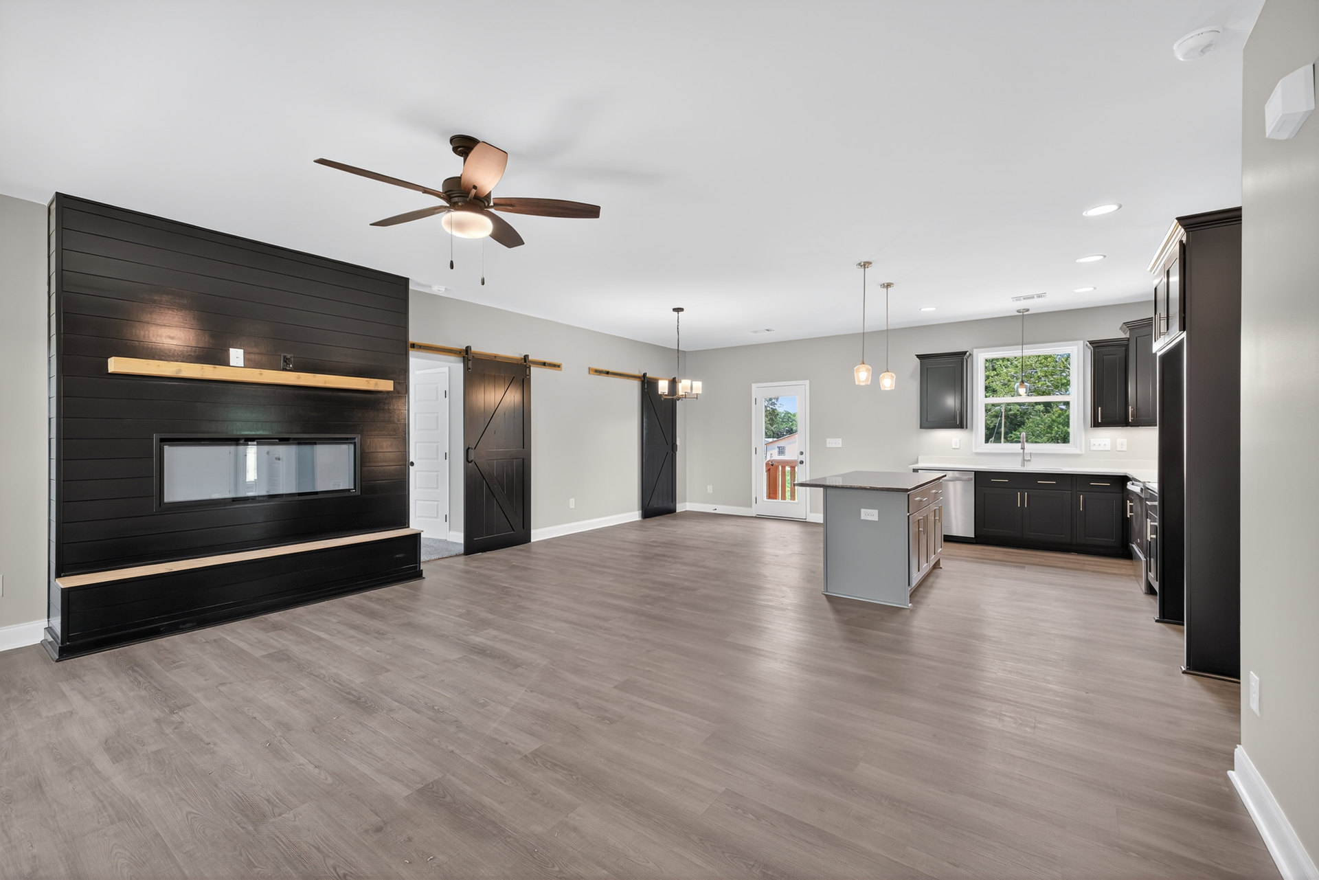 Spacious living area with wood flooring, black accent wall featuring a window, central fireplace, ceiling fan with light, and adjacent kitchen island with light switch