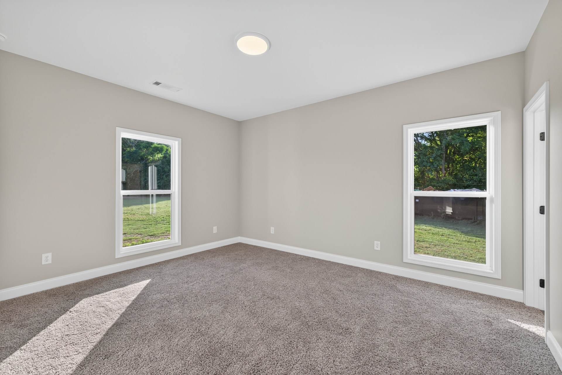 Carpeted room with large windows overlooking grass and trees, white walls, ceiling light fixture, and simple molding.