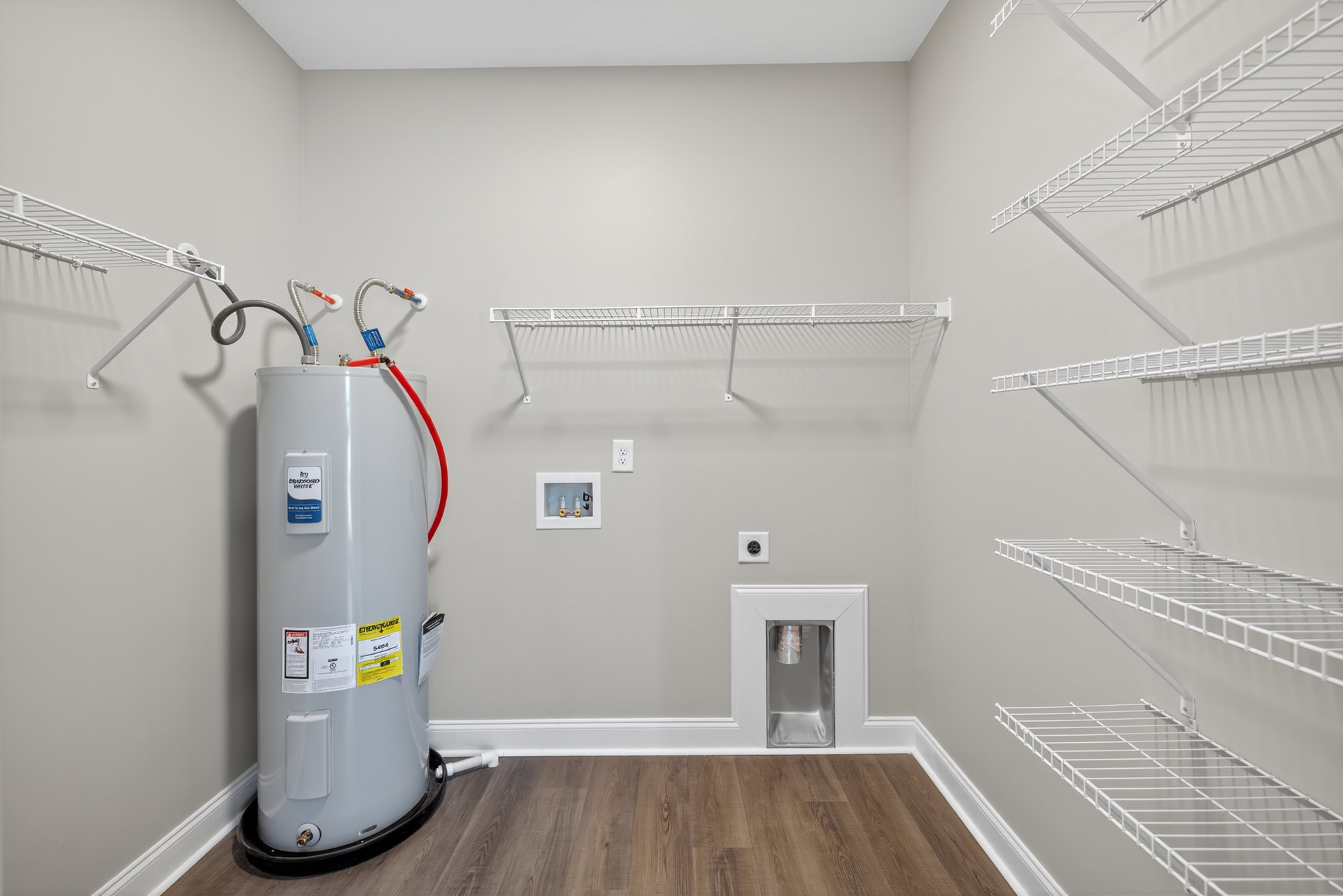 Utility room with white water heater featuring blue and yellow label, white shelving, metal pipe, and plaster walls