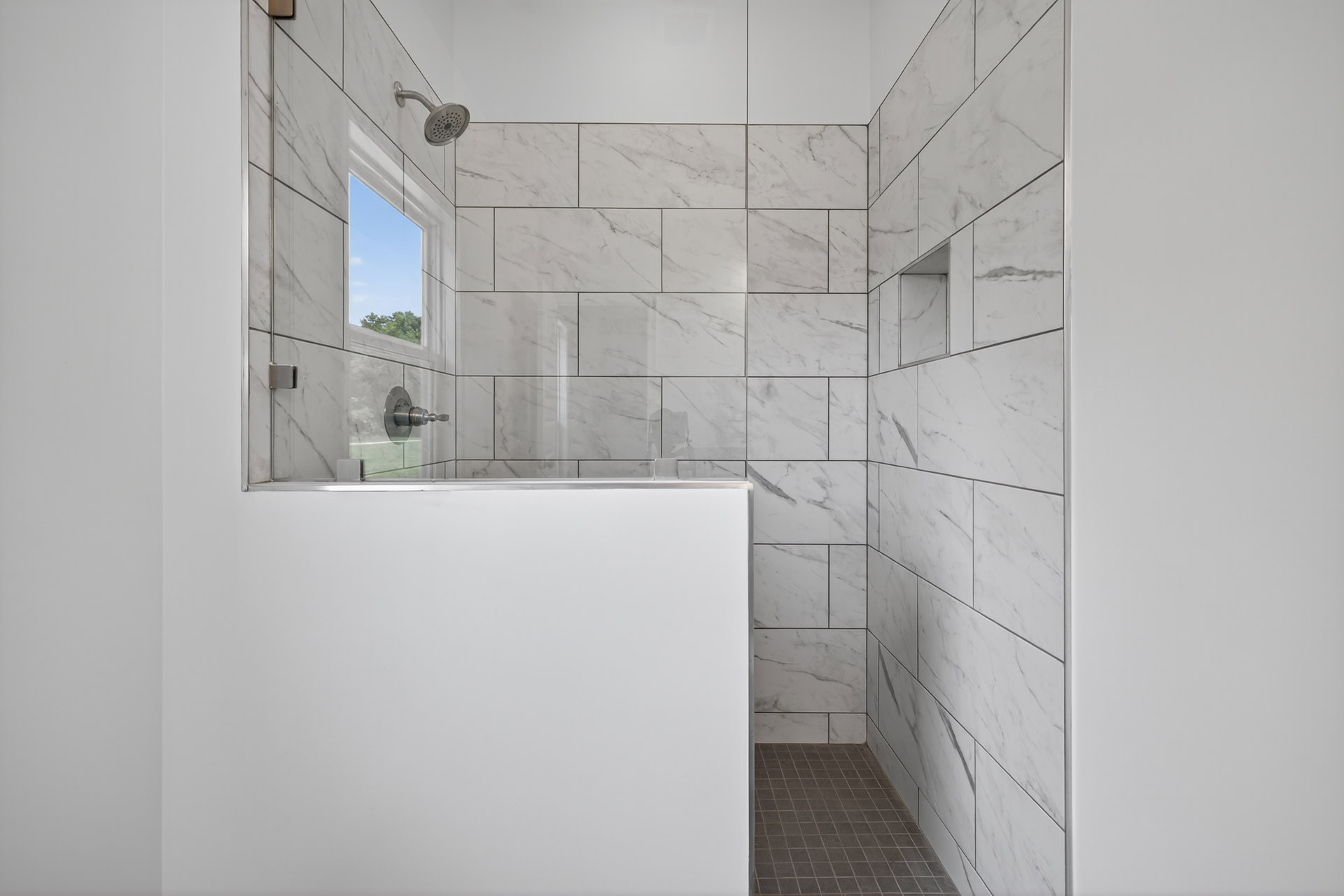 Walk-in shower featuring a patterned tile floor, white plaster walls, chrome shower head, and a window revealing blue sky.