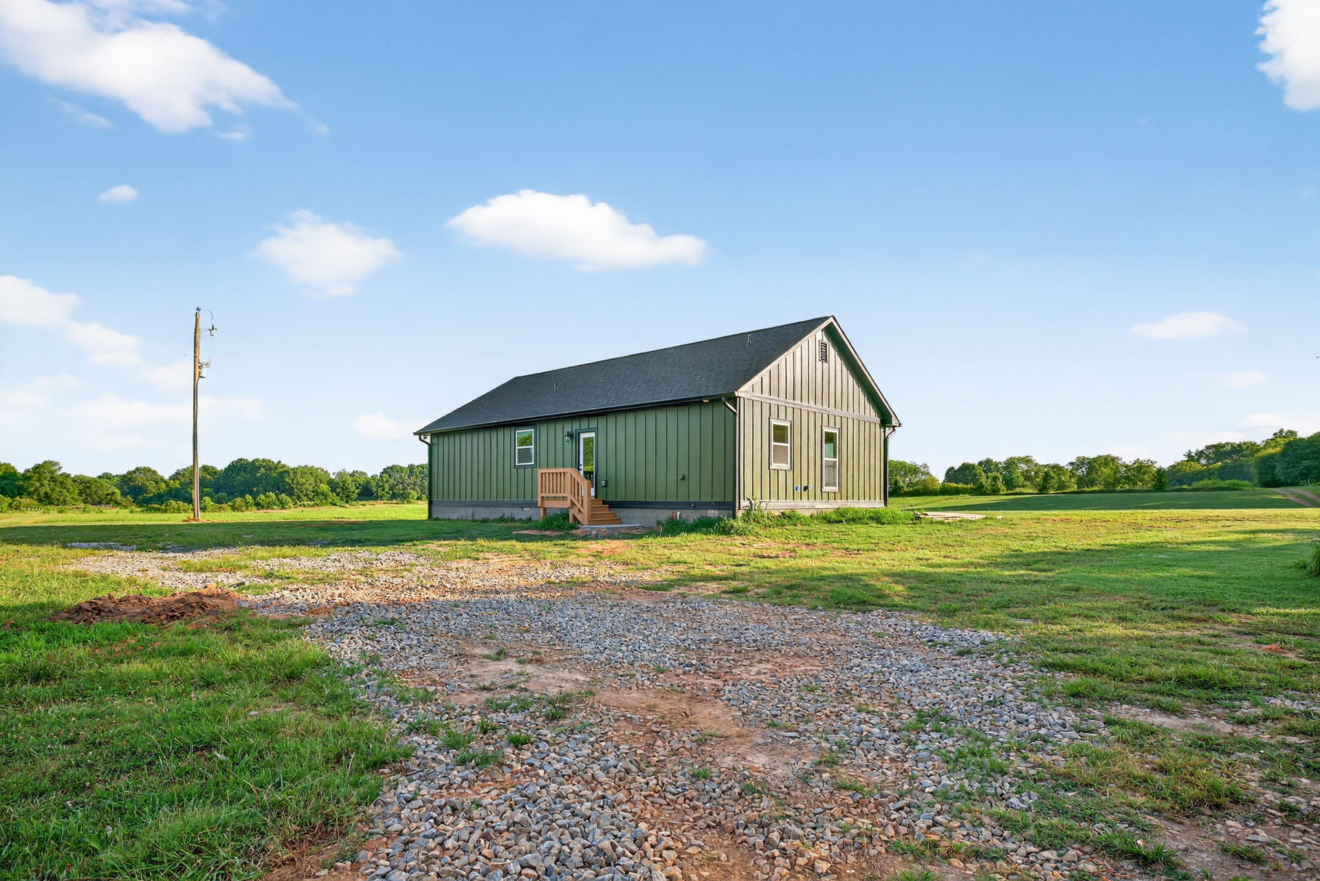 Green-painted house with wooden exterior staircase, gravel area bordered by grass, blue sky with scattered white clouds in rural landscape.