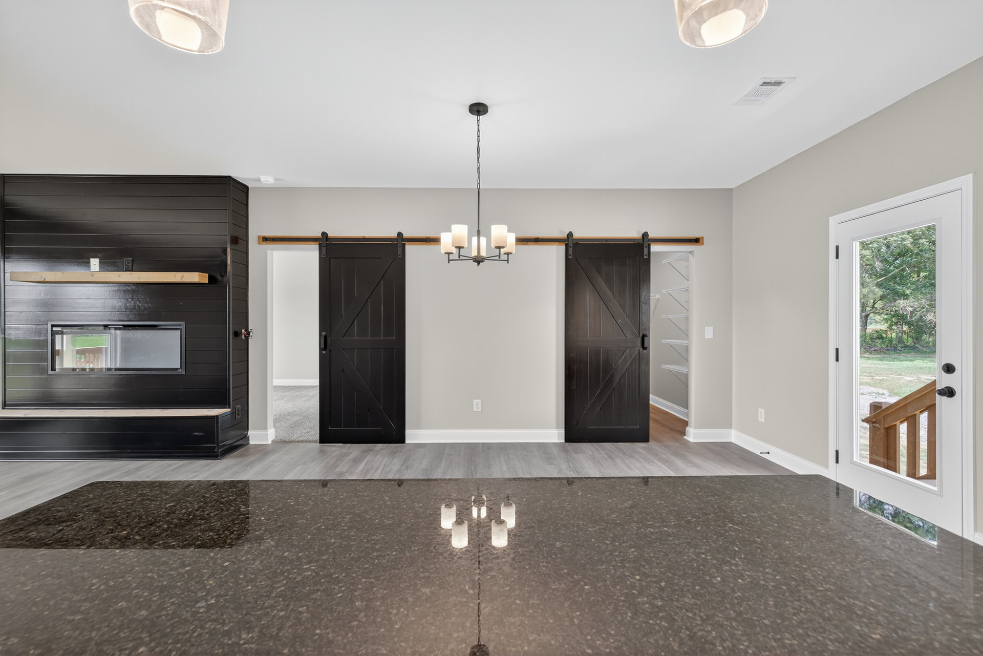 Modern living space featuring large sliding glass doors, a contemporary chandelier, black accent wall with window, light wood flooring, and minimalist cabinetry.