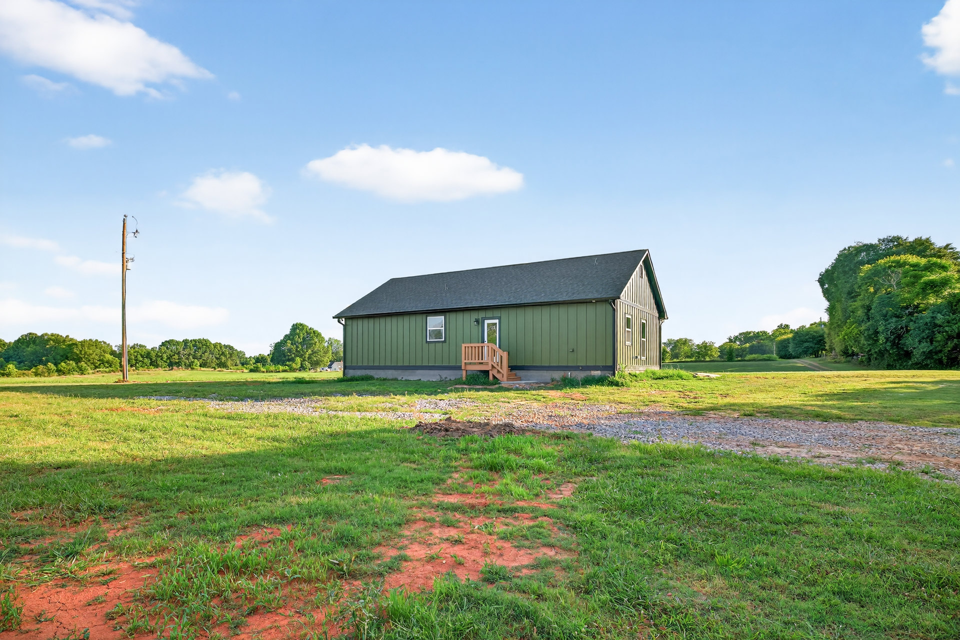 Green farmhouse with white roof, wooden porch and railing, surrounded by grassy field and trees under blue sky with clouds