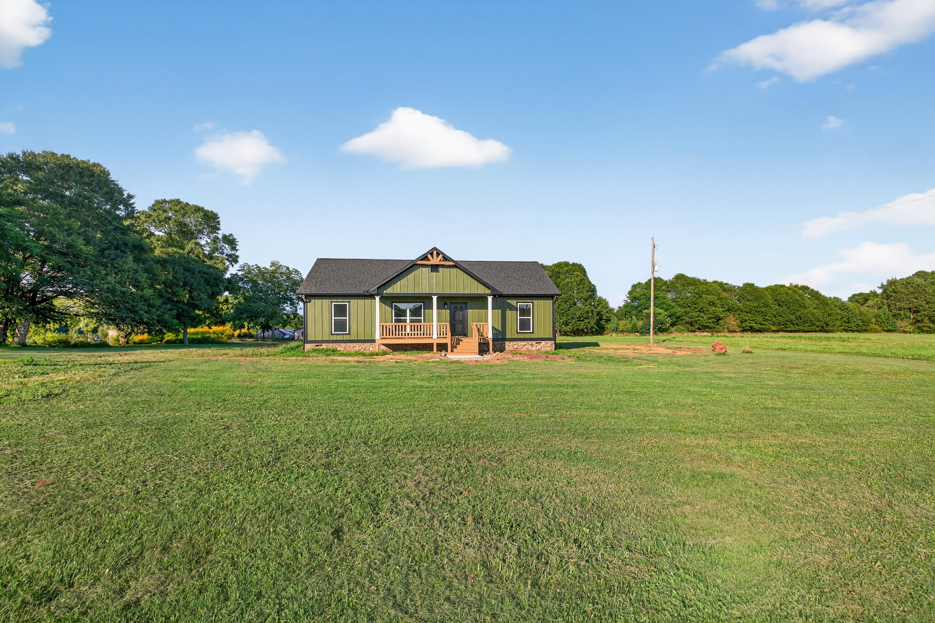 Green-painted farmhouse with wooden porch, large grassy lawn, mature trees, and white clouds in blue sky