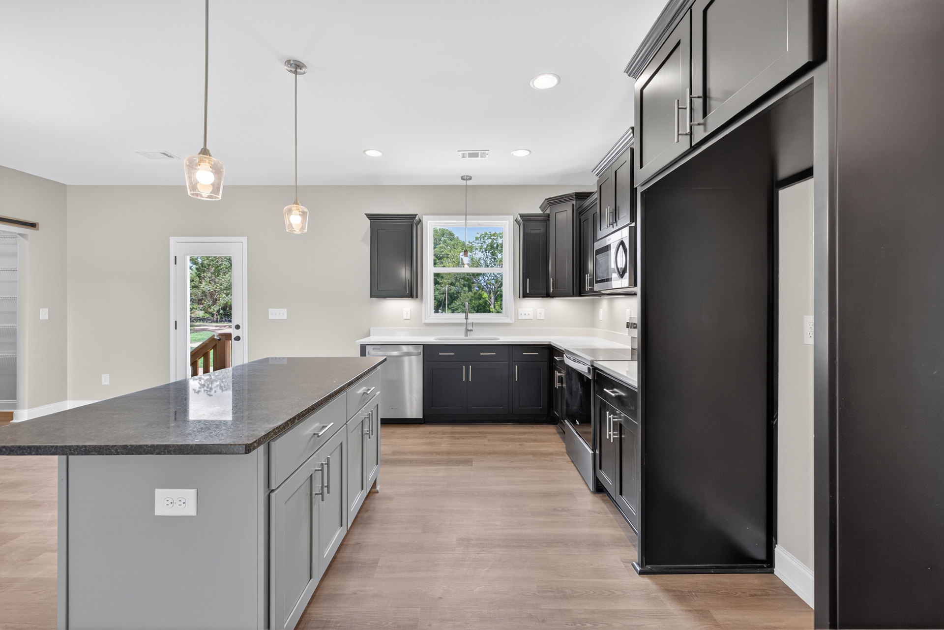 Black kitchen cabinets, wood flooring, granite-topped island, window with hanging rope, white wall with narrow trim, electrical outlet close-up, door opening to tree views