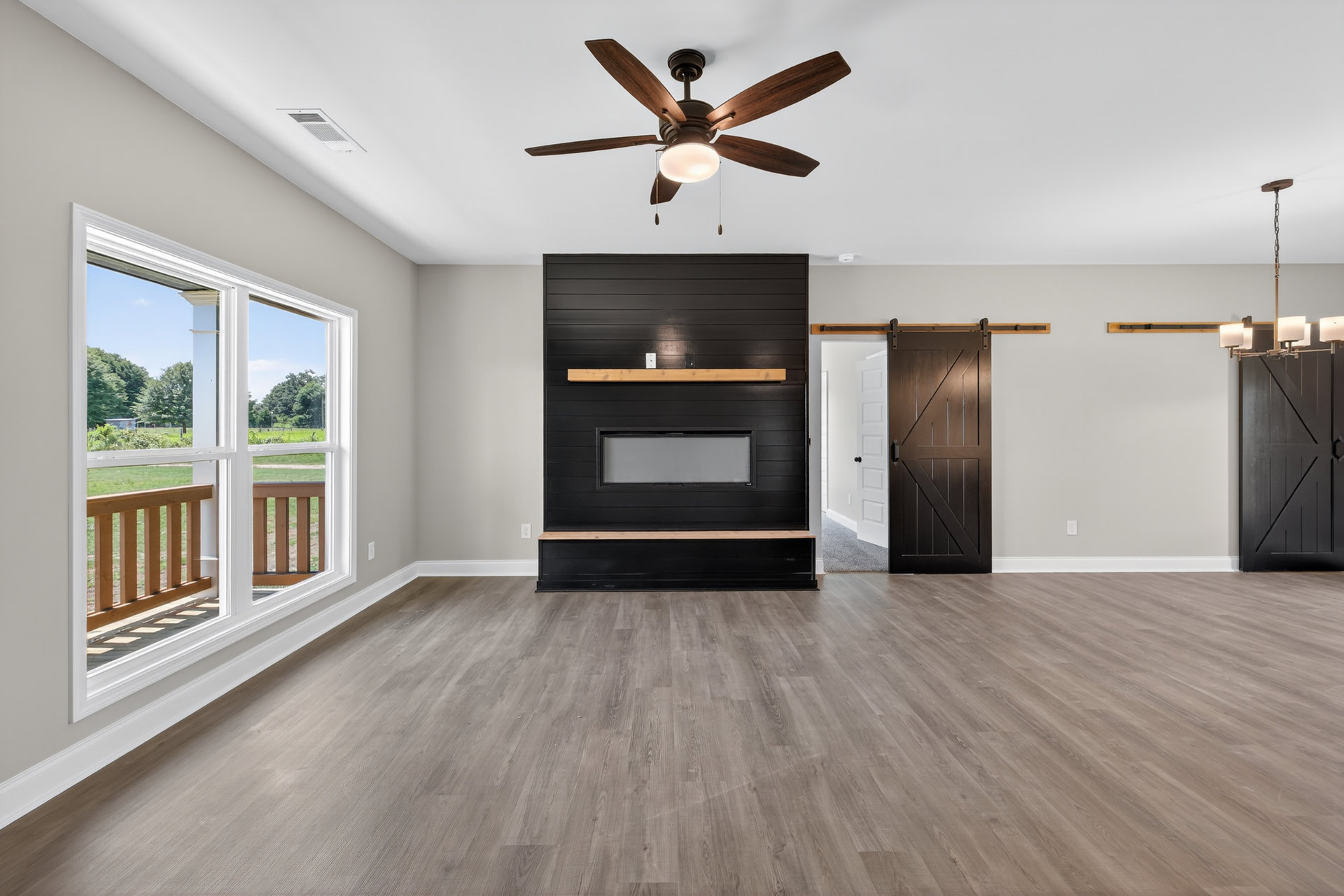 Living room with wood flooring, ceiling fan with light, built-in fireplace and shelf, upright piano, white door, large window overlooking yard
