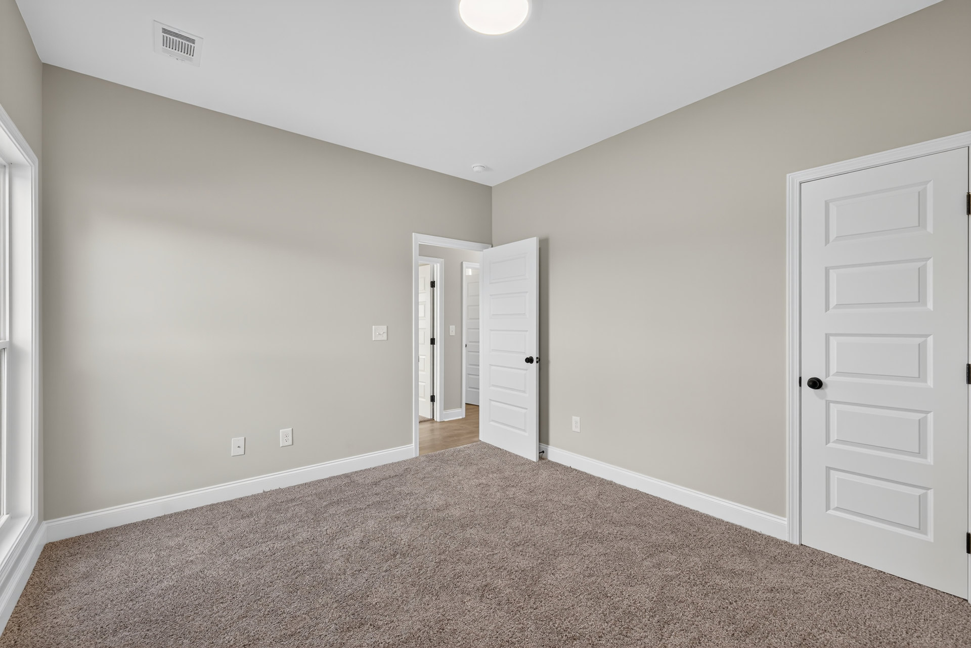 Bedroom with light gray carpet, white walls, two white doors featuring black handles, simple baseboard molding, and soft natural lighting.