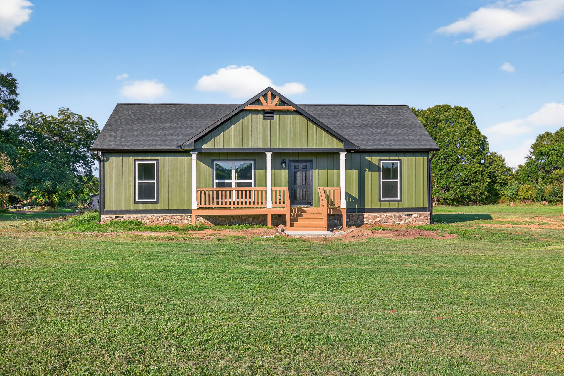 Green-painted cottage with white-framed windows, stone porch wall, blue curtain, and manicured lawn under a partly cloudy sky