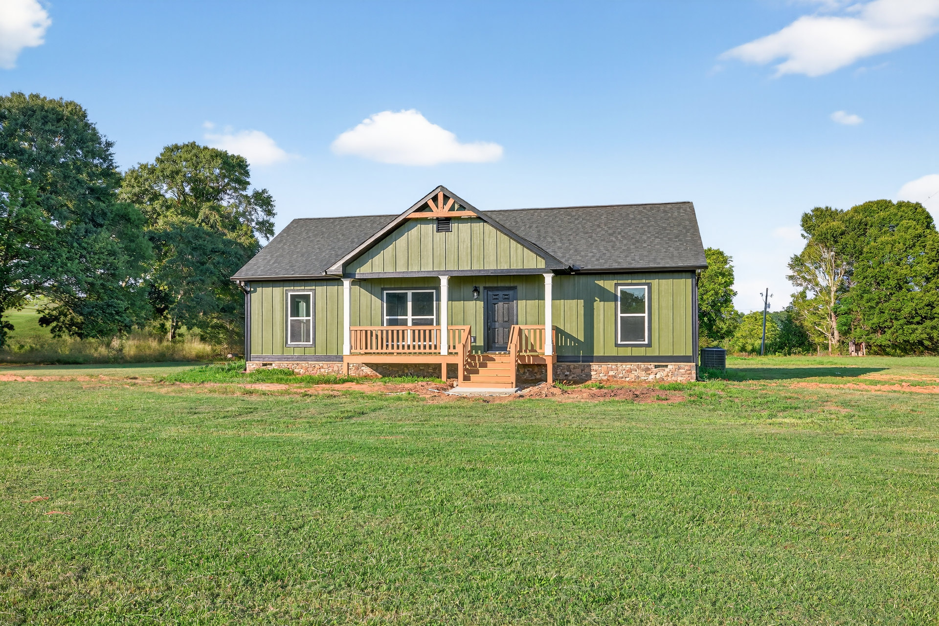 Green-painted farmhouse with white-trimmed windows, wooden porch featuring stone wall accents, expansive grassy lawn, and mature leafy tree under a partly cloudy sky