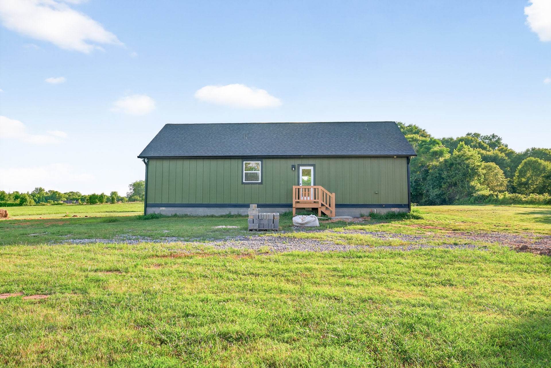Green-painted house with white-framed windows, covered porch, white door with glass inset, grassy lawn featuring a wooden chair and stacked bricks, surrounded by open field and