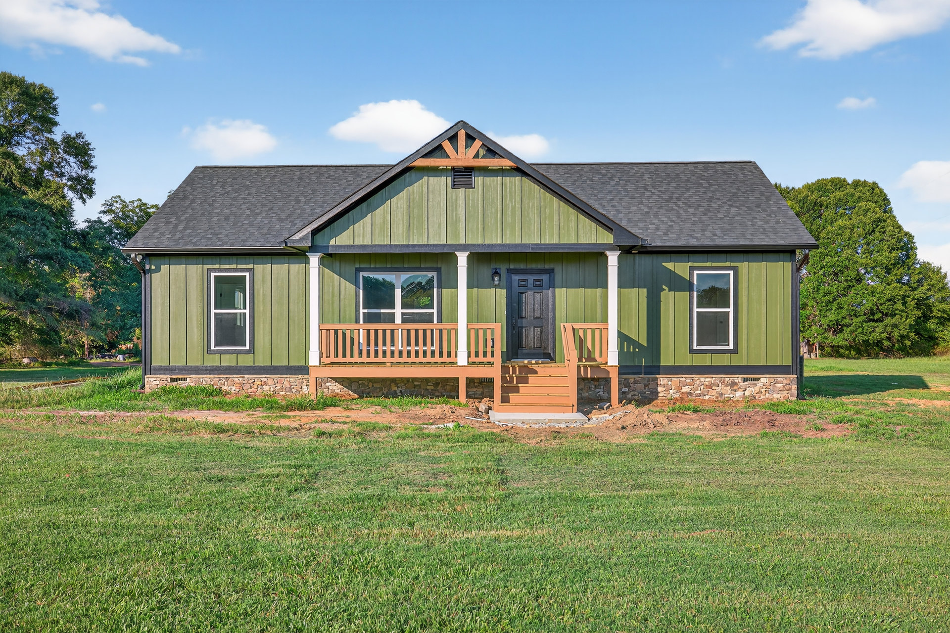 Green siding house with white trim, covered porch featuring stone wall accents, manicured lawn, large windows reflecting nearby trees, gabled roof with mature tree in background.