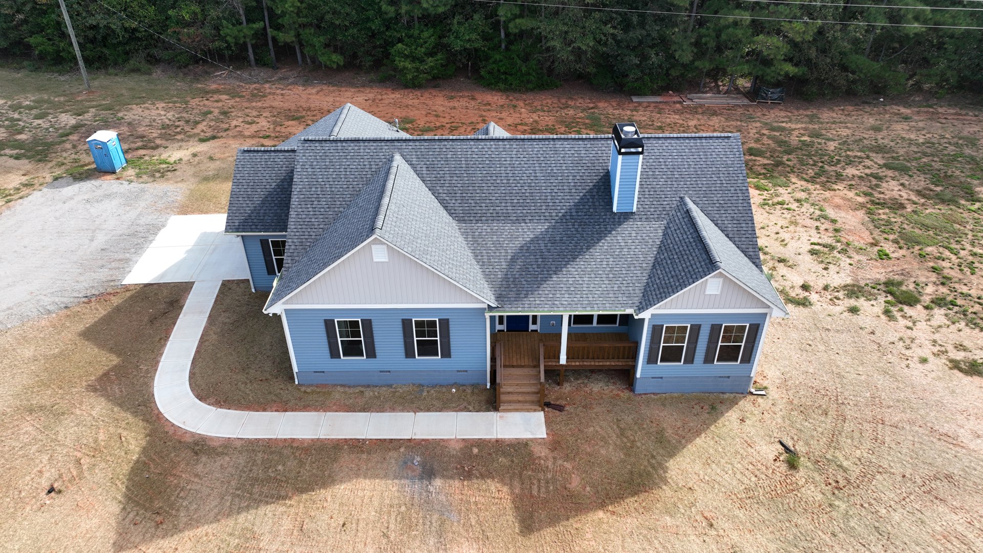 Two-story house with white-framed windows, metal chimney, wooden stairs leading to entry, paved driveway, mature trees, and blue portable toilet on grassy lawn