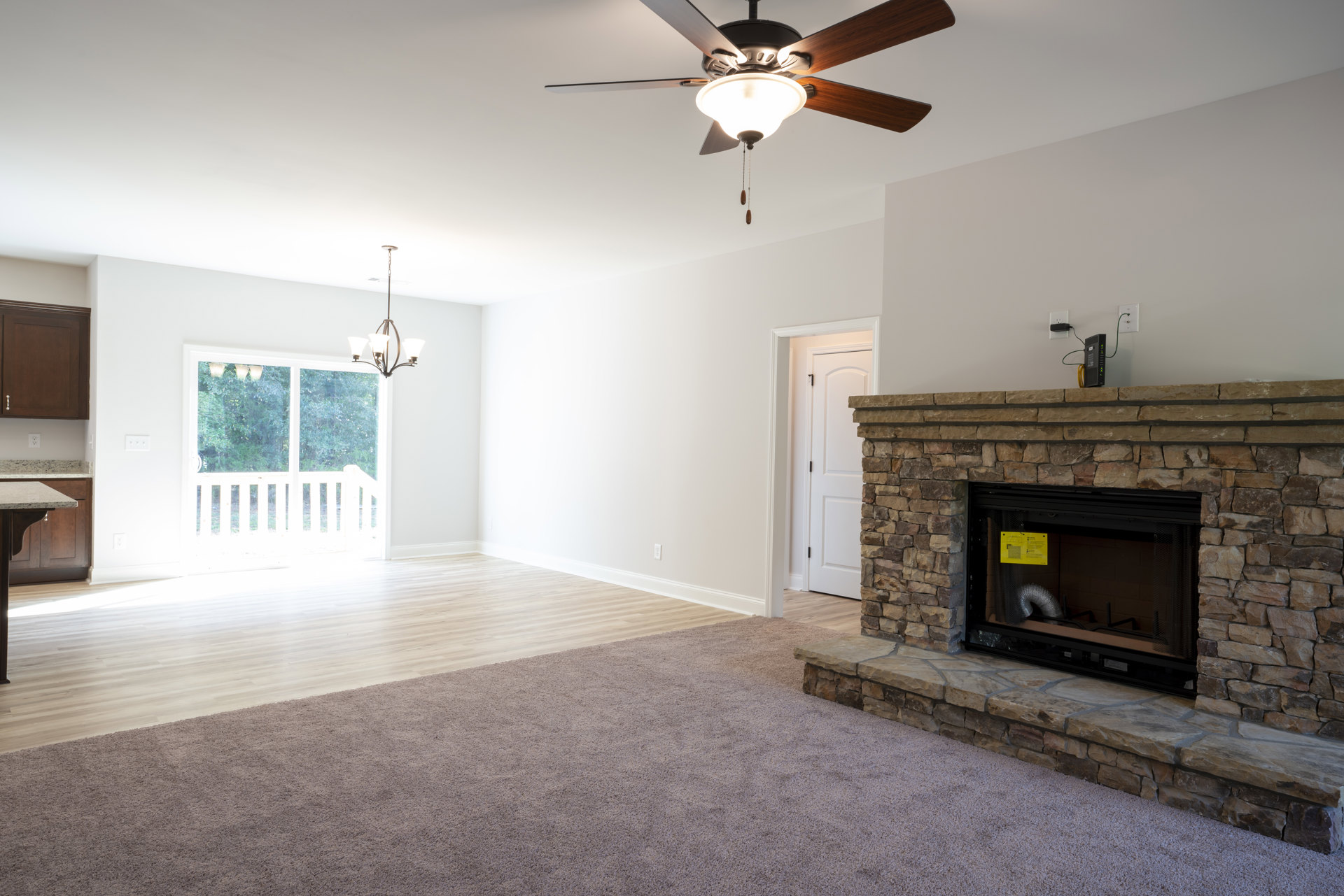 Living room with wood flooring, stone fireplace featuring a metal flue, ceiling fan with light fixture, white-framed window with railing, and paneled door.