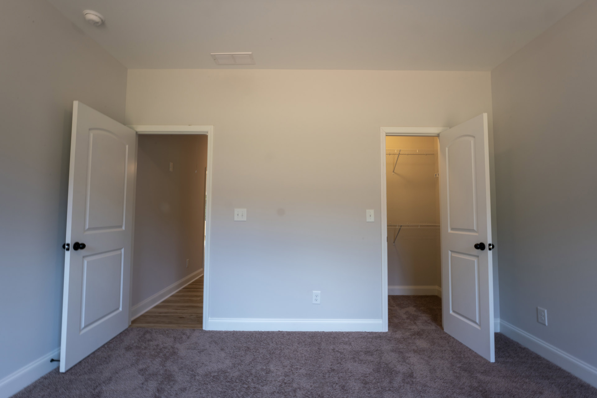 Bedroom with two open white doors featuring black knobs, beige carpet flooring, white walls, and a closet with a built-in shelf illuminated by natural light.