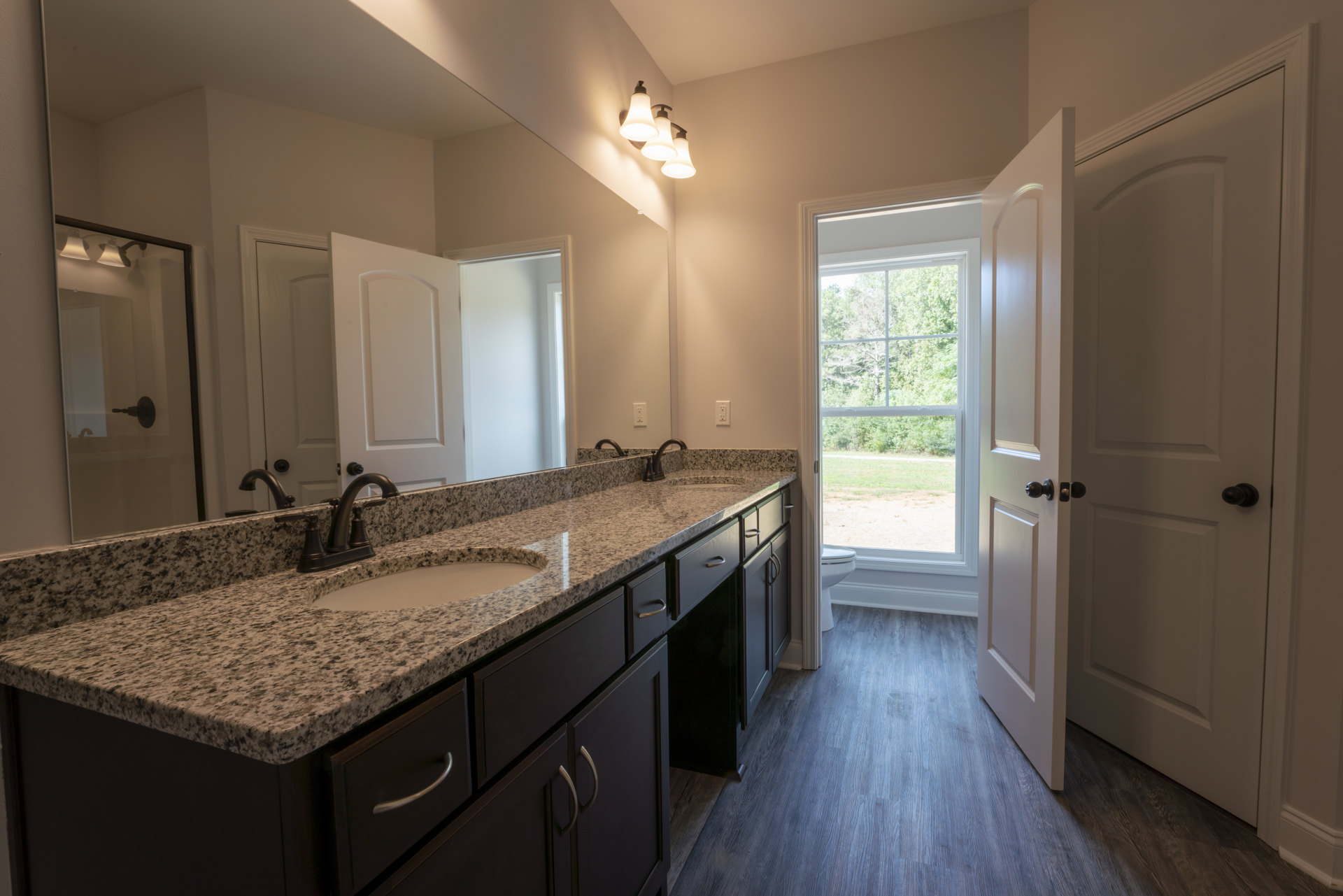 Bathroom with marble countertop, undermount sink, chrome faucet, three-light fixture above large mirror, white cabinetry with black knobs, door, window showing trees outside