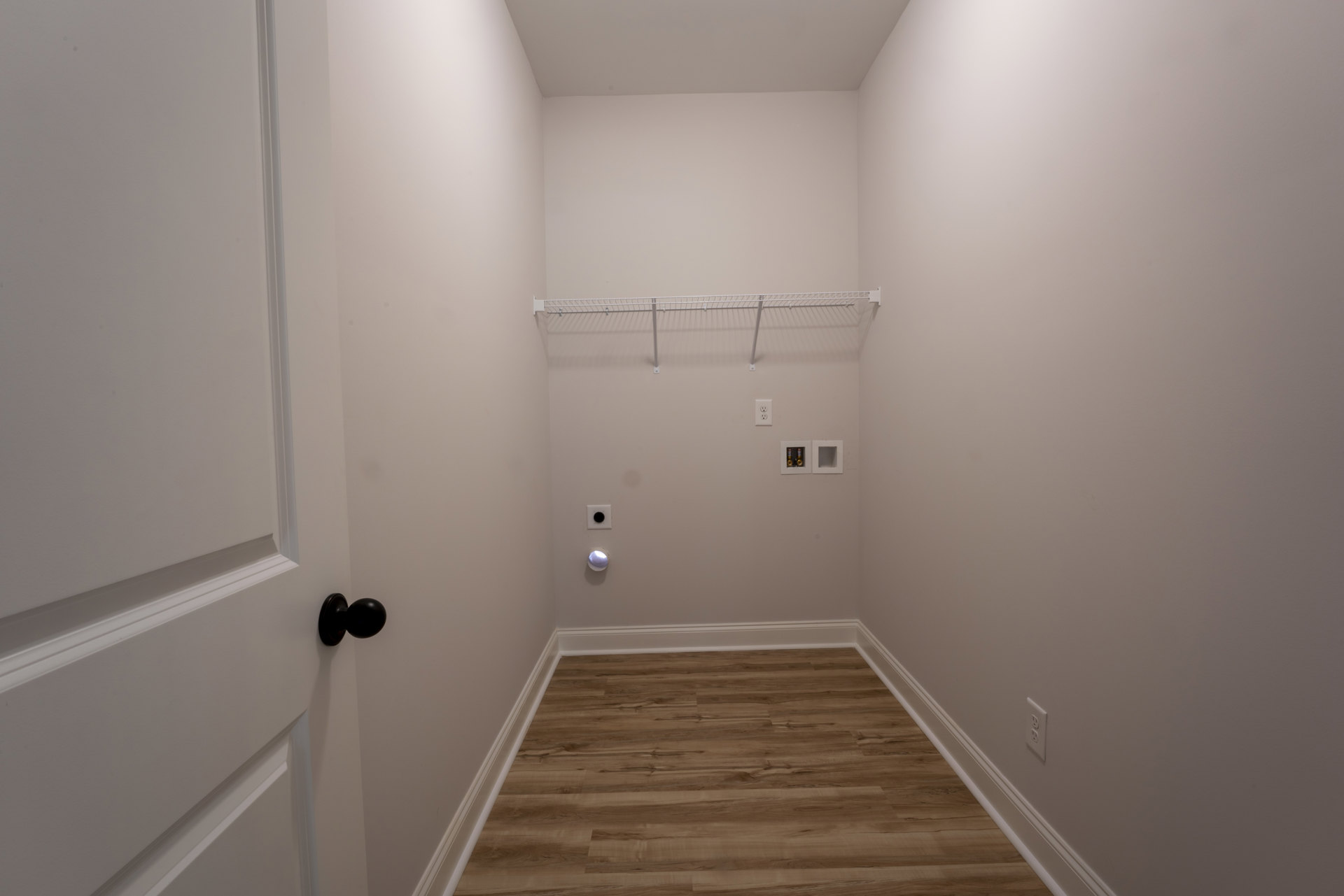 Hallway with light wood flooring, white baseboards, white door featuring a black knob, white metal rack with hooks mounted on the wall, and gold accents inside a white square