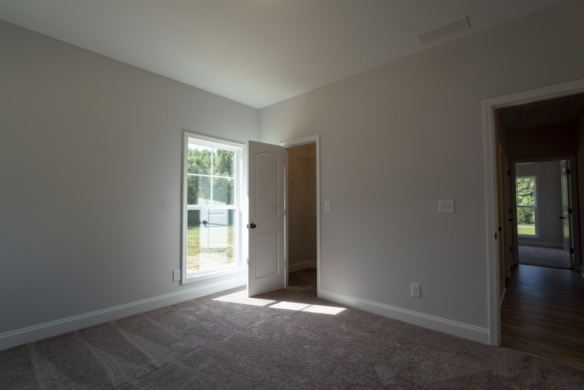Open white door and window in a room with dark wood laminate flooring, sunlight streaming onto the floor, white plaster walls, and view of greenery outside.
