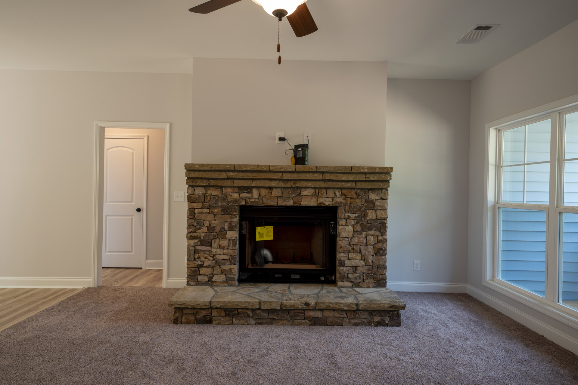 Stone fireplace with yellow sign on mantel, carpeted floor, white door with black knob, multiple windows, ceiling fan above.