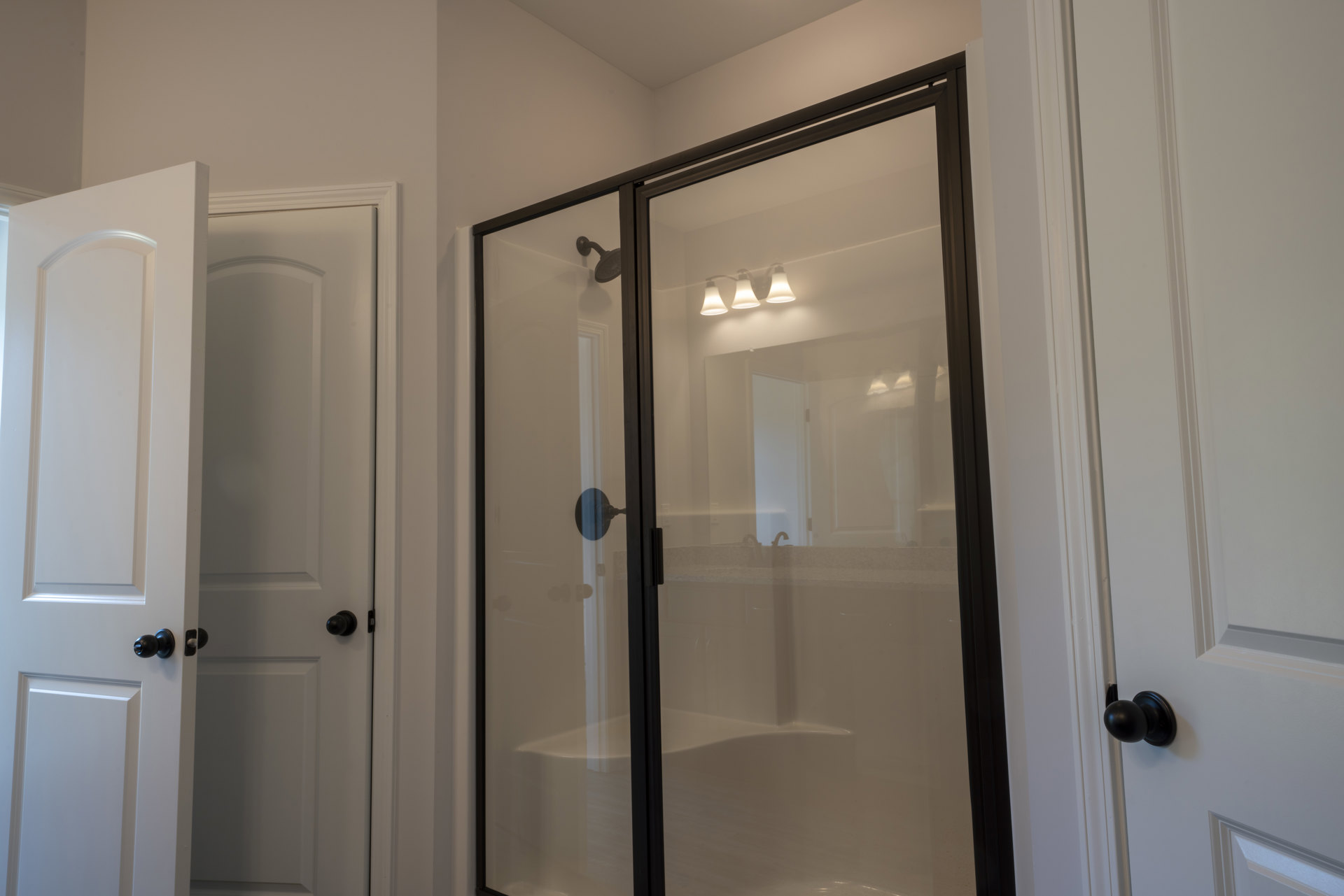 Frameless glass shower enclosure with chrome hardware, white tile walls, and a modern light fixture with a white shade above a rectangular mirror; black door knob on a white